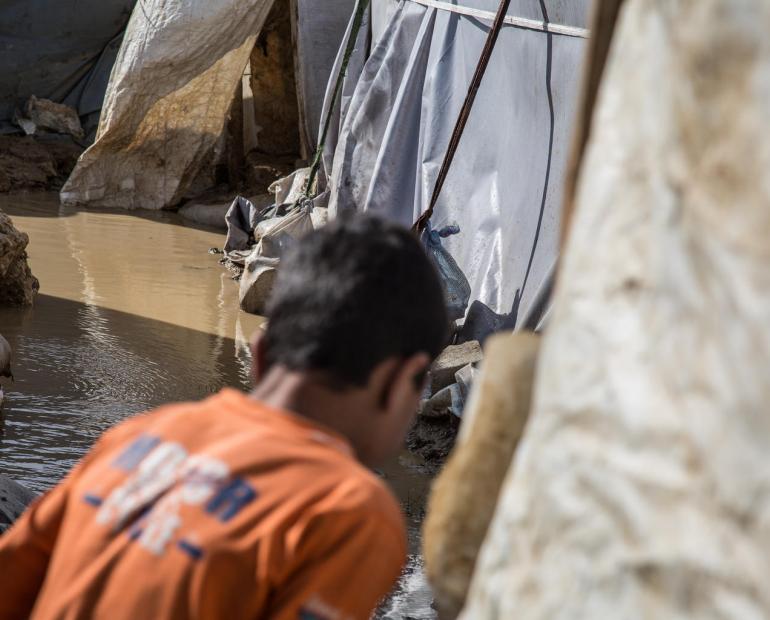 A Syrian refugee child, his back to the viewer, checks the water canal surrounding his  tent in an informal settlement in the Bekaa Valley, Lebanon, 2018.