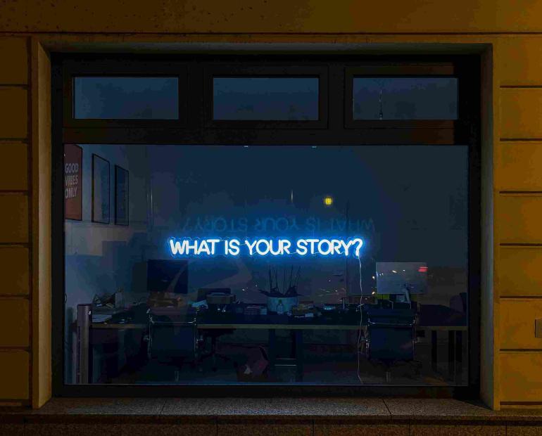 A blue neon sign that says "What is your story?" is on the wall of a room, behind a desk with objects on top.
