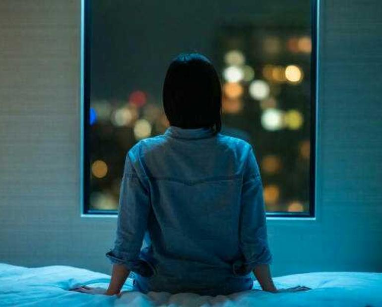 A girl sitting alone on a bed looking at a window 