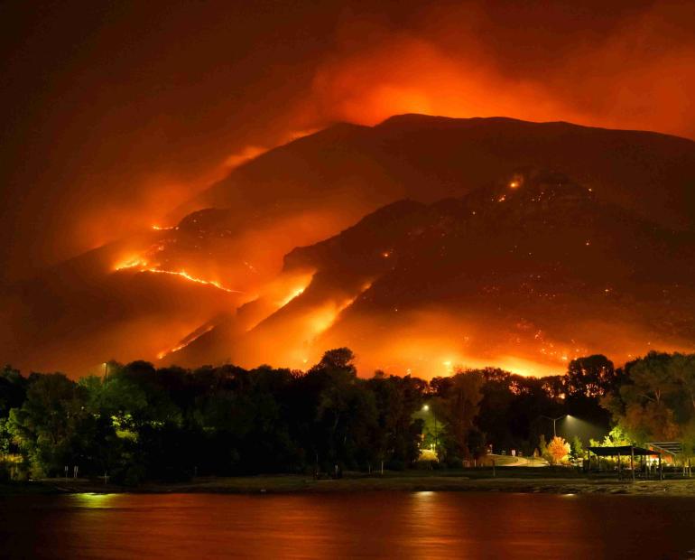 A picture of a mountain on fire at night, the flames illuminate the tress and lake.