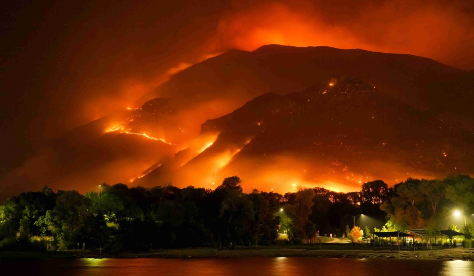 A picture of a mountain on fire at night, the flames illuminate the tress and lake.