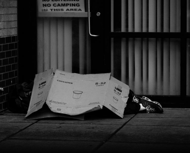 A black & white photo of a person laying under a cardboard box on the streets