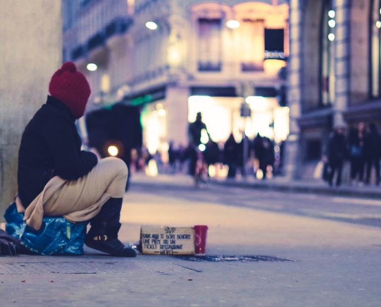 Person sitting on the street covered with a blanket near a cardboard sign asking for help