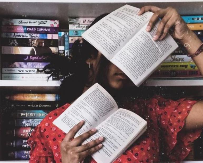 Person, the writer of the piece, is holding two books in front of her face, which cover her eyes. Person is standing in front of a bookshelf with a large number of books behind her.