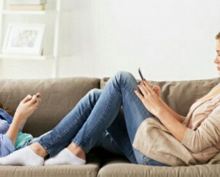 A mother and her teenage girl sitting face to face on a sofa. Both of them are  using their smartphone. 