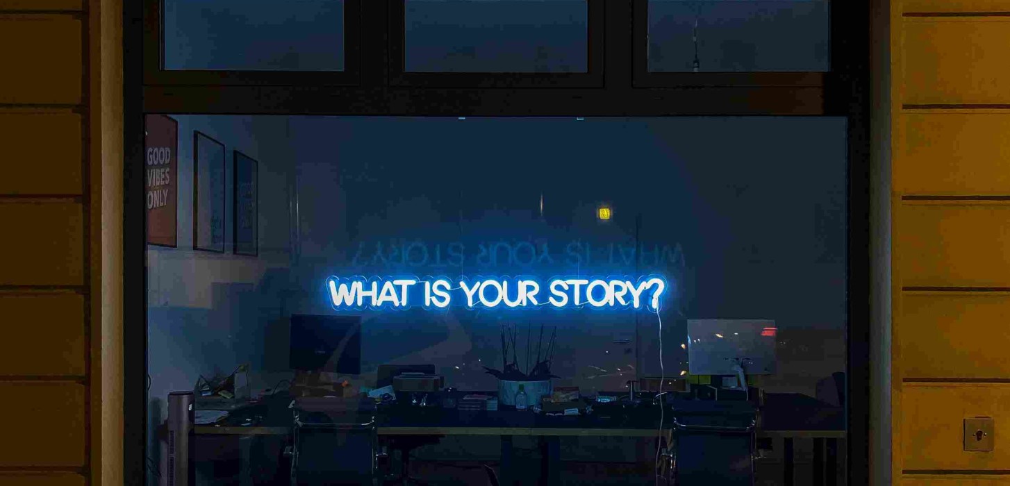 A blue neon sign that says "What is your story?" is on the wall of a room, behind a desk with objects on top.