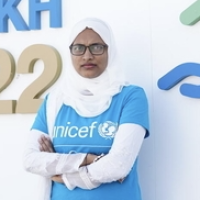 Farzana wears a UNICEF cyan t-shirt and stands before a sign that says COP 27