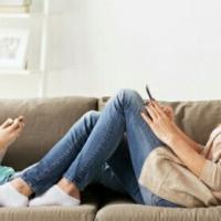 A mother and her teenage girl sitting face to face on a sofa. Both of them are  using their smartphone. 